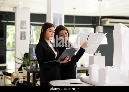 Focused women study architectural blueprints and analyze design elements for upcoming project. Female designer prepare essential online documentation needed for accurate construction site execution. Stock Photo