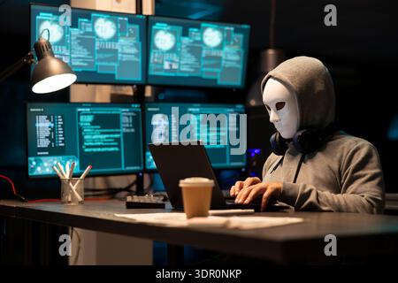 Masked male individual works on laptop at night, attempting to breach corporate systems. Anonymous hooded man appears to hack software company, representing cybercrime and digital security threats. Stock Photo