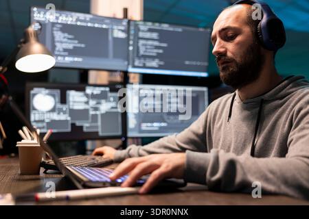 Bearded man wearing headphones concentrates on laptop while glowing monitors display technical interface. Freelance programmer listens to music and works on software development code in office. Stock Photo