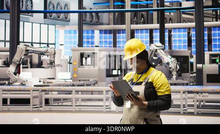 Woman working in solar panels plant uses tablet to oversee machinery systems. African american photovoltaics plant employee uses software on device to maintain operations, camera A Stock Photo