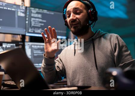Caucasian IT specialist wearing headset speaks during virtual meeting at desk. Multiple digital screens show coding software as programmer supports clients and collaborates on technical solutions. Stock Photo