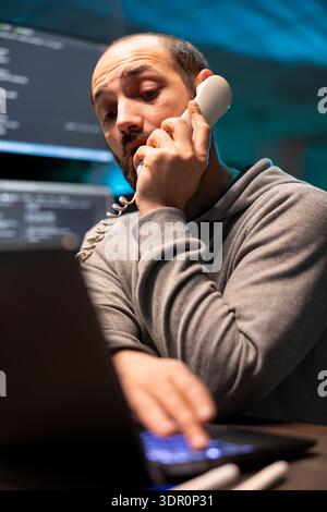 Busy male programmer speaks on desk phone, juggling tech support and website tasks. Caucasian developer works in fast paced workflow, communicating with clients and solving cybersecurity challenges. Stock Photo