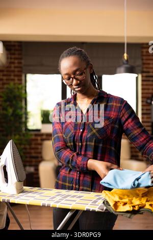 African american female individual organizing folded shirts on ironing board after ironing. Black woman homeowner manages pile of freshly pressed clothes, completing productive home laundry routine. Stock Photo