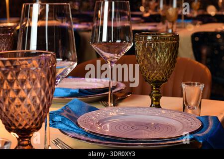 Closeup elegantly set restaurant table featuring decorative colored goblets, wine glasses, plates, folded blue napkins. Warm ambient lighting creates Stock Photo