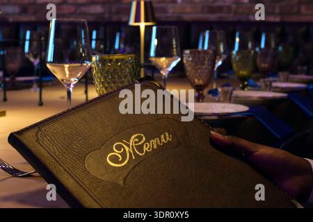 Closeup person hand holding leather bound menu while seated at elegantly set restaurant table. Fine dining atmosphere with table setting, glassware, a Stock Photo