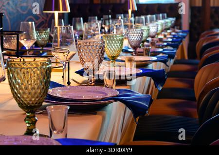 Elegant banquet or restaurant table with colored goblets, wine glasses, blue napkins, and white plates. Warm lighting creates a formal dining atmosphe Stock Photo
