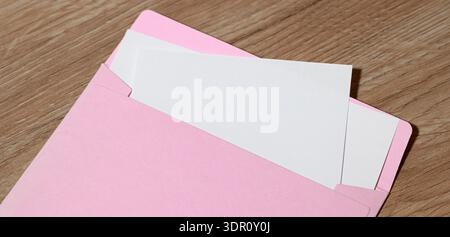 A close-up shot shows a pastel pink envelope slightly open, revealing blank white paper inserts against a textured wooden background Stock Photo