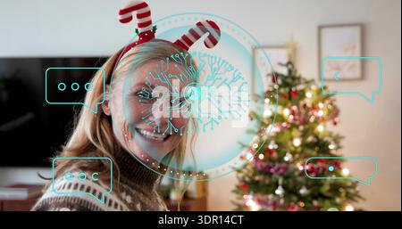 Smiling woman in patterned-sweater, candy-cane-headband facing camera at home with Xmas tree TV AR Stock Photo