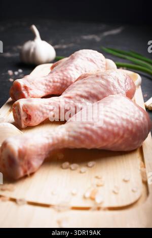 Top view of three raw chicken drumsticks on a wooden cutting board with garlic, green onions, and pink salt, set against a dark marble background for Stock Photo
