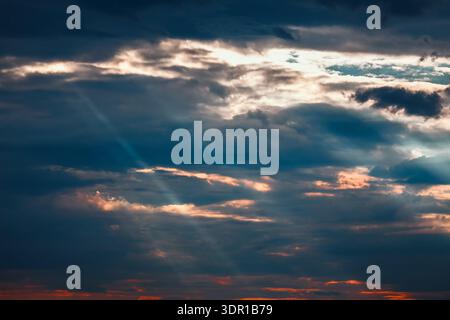 Dramatic crepuscular rays piercing through dark, layered stratocumulus clouds at sunset. Sun is setting, casting a warm glow on the clouds in the sky Stock Photo