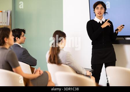 Confident young malaysian female student with remote control confidently took stage, ready to deliver impactful presentation to audience in classroom Stock Photo