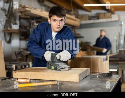 Portrait of young man carpenter using electric sanding machine to polish wood barin workshop Stock Photo