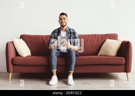 Young Man Sits on Red Sofa Holding Tablet and Smiling in a Modern Living Room Setting Stock Photo