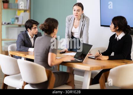 Confident young businesswoman standing near table where sitting colleagues talking and discussing ideas in office Stock Photo