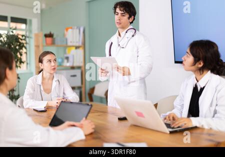 Confident young guy medical student standing near table where sitting interns talking and discussing ideas during meeting in office Stock Photo