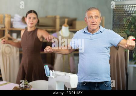 Elderly man and young woman doing warm-up exercises in sewing workshop Stock Photo