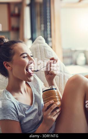 Woman, happiness and eating peanut butter in home with jar, taste and vegan diet for wellness in lounge. Person, food nutrition or spoon with nut Stock Photo