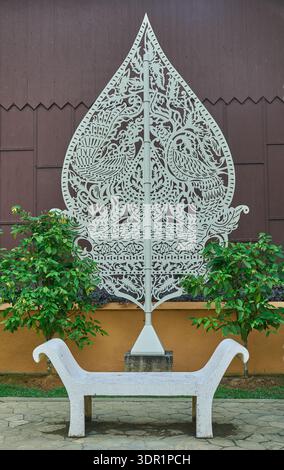 Intricately carved leaf-shaped metal sculpture mounted behind a white curved concrete bench in a landscaped garden. Stock Photo