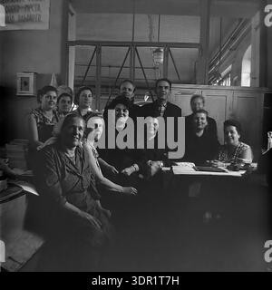 Slavyansk, Ukraine, USSR. 1960s. A large group portrait of a production team posing together at the Slavyansk Pencil Factory. The black and white archival photo shows a mixed collective of manual workers in heavy overalls and administrative staff in casual clothing. They are gathered around a desk with papers and an inkstand in a workshop office area. Two men stand in the back row among the women. A motivational slogan is visible on the wall. The image captures the unity, camaraderie, and professional pride of the workforce in the peaceful Donbas region. Stock Photo