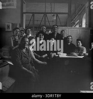 Slavyansk, Ukraine, USSR. 1960s. A large group portrait of a production team posing together at the Slavyansk Pencil Factory. The black and white archival photo shows a mixed collective of manual workers in heavy overalls and administrative staff in casual clothing. They are gathered around a desk with papers and an inkstand in a workshop office area. Two men stand in the back row among the women. A motivational slogan is visible on the wall. The image captures the unity, camaraderie, and professional pride of the workforce in the peaceful Donbas region. Stock Photo