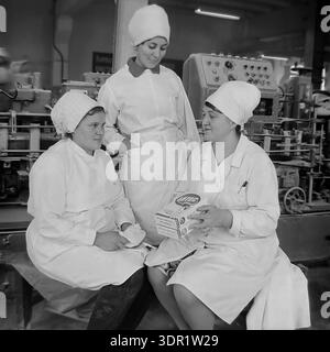 Slavyansk, Ukraine, USSR. 1960s. Three female factory workers in white lab coats and head covers discuss consumer goods production at the Slavyansk Oil and Fat Plant. The black and white archival photo captures employees sitting next to a complex packaging machine while inspecting "Lotos" (Lotus) detergent boxes. One woman holds the product while her colleagues look on. The background features technical control panels and industrial equipment. This image highlights female employment, chemical industry development, and the peaceful atmosphere of the Donbas region. Stock Photo