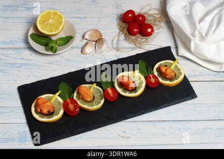 Mussels tapas appetizers arranged on a black slate board, garnished with lemon, mint leaves, and cherry tomato halves Stock Photo