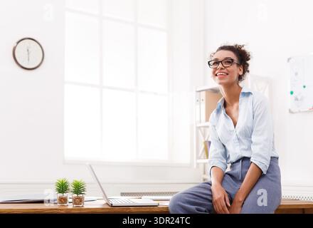 Young Woman Sitting on Desk Showing Smile in Bright Office Space Stock Photo