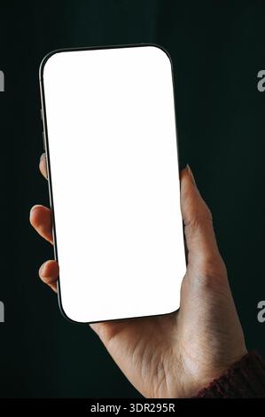 Close-up of a hand holding a modern smartphone with blank white screen against dark background. Ideal mockup for mobile app design, digital advertisin Stock Photo