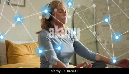 Meditating senior woman wearing headset and leggings on bed at home mustard pillow glowing network Stock Photo