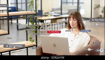 Working woman wearing white shirt using silver laptop in office lounge with shelving, floating UI Stock Photo
