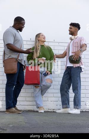Three friends chatting while standing in front of white painted brick wall holding red shopping bag Stock Photo