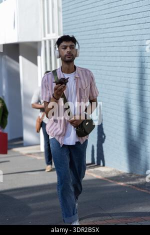 Asian man walking sidewalk by blue brick wall wearing white headphones with green bag holding phone Stock Photo