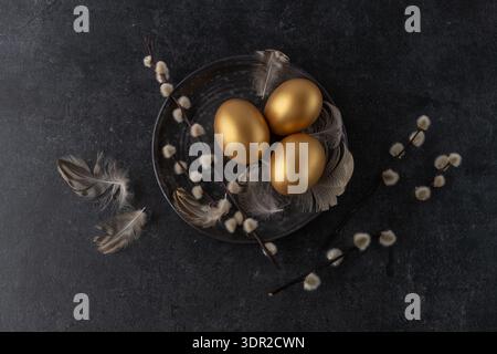 Easter painted golden natural chicken eggs with willow branches and bird feathers on vintage dark grey plate on black stone background with copy space Stock Photo