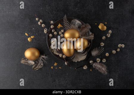 Easter golden painted natural chicken eggs with willow branches and  fluffy bird feathers on vintage dark grey plate on black stone background. Happy Stock Photo