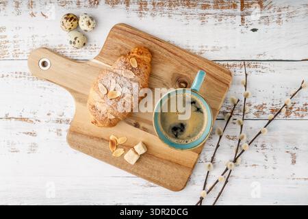 Fresh baked almond nuts breakfast croissant and hot coffee vintage turquoise cup over cutting board with willow branches and quail eggs on rustic wood Stock Photo