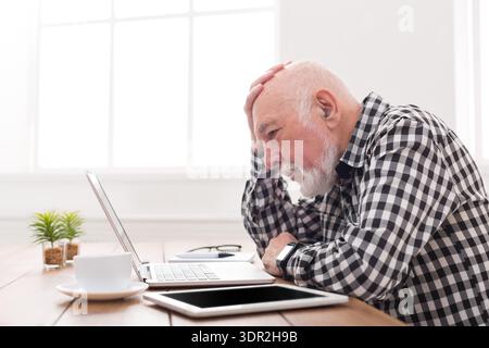 Frustrated Older Man Working on Laptop While Sitting at Table in Bright Room Stock Photo