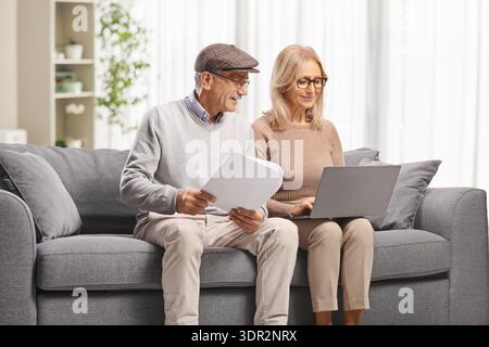 Mature man reading a document and woman using a laptop computer seated on a sofa Stock Photo