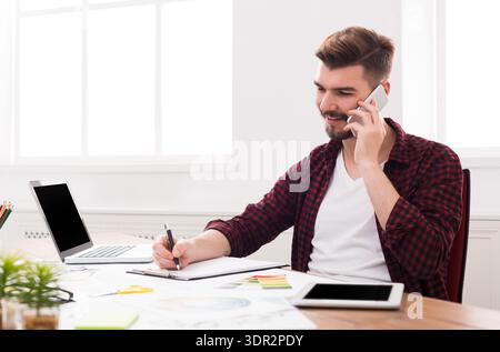 Young Man Working at a Desk While Talking on the Phone and Taking Notes Stock Photo