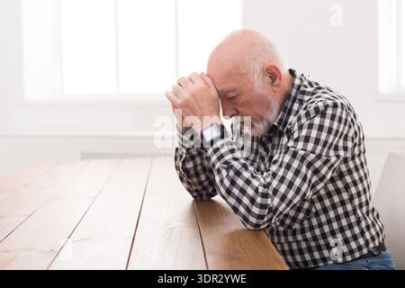 Elderly Man Sits at Table With Hands Clasped, Deep in Thought in a Light Room Stock Photo