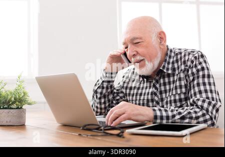 Senior Man Using Phone and Laptop While Sitting at Table With Tablet Nearby Stock Photo