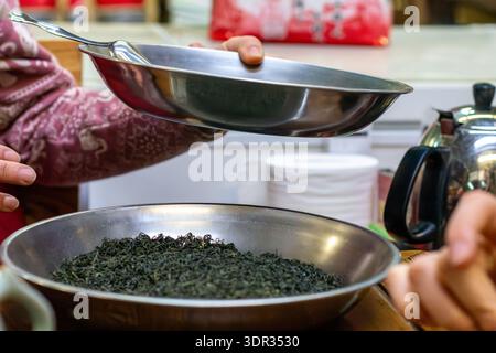Hands holding metal pan and strainer over large bowl of dried tea leaves in a kitchen, close-up. Concept of sorting, processing, and preparing loose-l Stock Photo
