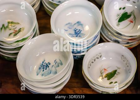 Stacks of small white porcelain tea cups with hand-painted bird and landscape motifs, arranged on a wooden table in a shop display. Stock Photo