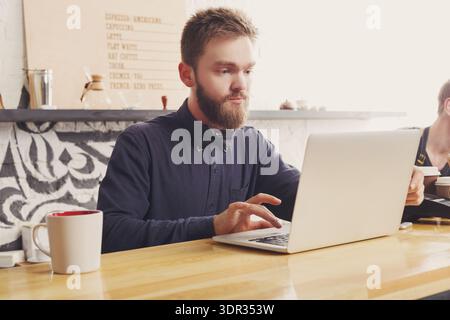 Young Man Works on Laptop at Coffee Shop During Busy Day Stock Photo