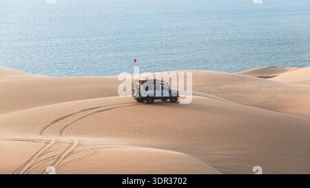 View of a vehicle atop rippling dunes, tire tracks etched in the sand, against the backdrop of the vast, tranquil ocean, Walvis Bay, Erongo Region, Na Stock Photo