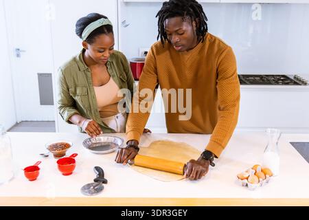 African American couple making dough in kitchen using rolling pin, pie tin, woman wearing headband Stock Photo