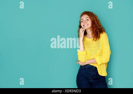 Young Woman Stands Against a Light Blue Wall While Smiling and Holding Her Hand on Her Chin Stock Photo