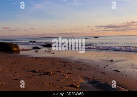 Gentle ocean waves rolling onto sandy beach at sunset with warm golden light, scattered stones, soft clouds and peaceful horizon over calm sea. Stock Photo