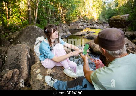 Family hikers using navigation app on smartphone in forest. Daughter showing map to father during trekking. Technology in nature and travel concept. Stock Photo