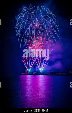 Stunnig image captures a vibrant fireworks display lighting up the night sky, with vibrant hues of blue, pink and purple reflecting on the calm waters Stock Photo