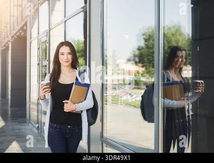 Student Standing Near Glass Building With Books and Phone at the University Campus in the Morning Stock Photo